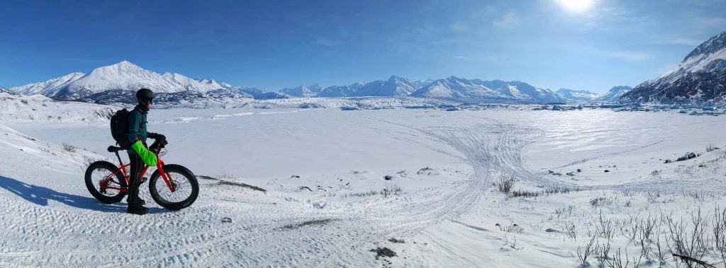 cyclist taking a break on a snow-covered landscape with the scenic, icy terrain of Knik Glacier