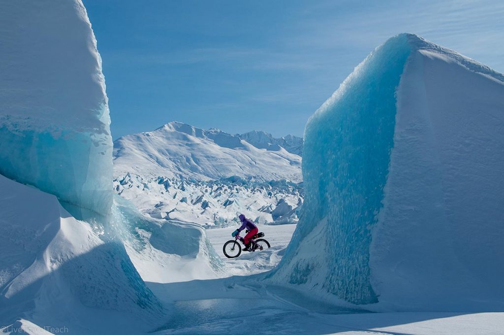 person riding a fat bike between massive blue icebergs on the frozen surface of the Knik Glacier in Alaska
