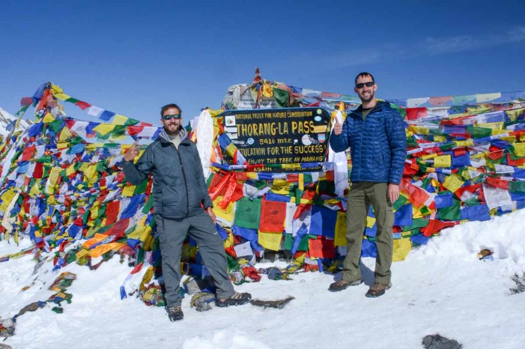 men standing by Thorong La Pass sign surrounded by colorful prayer flags